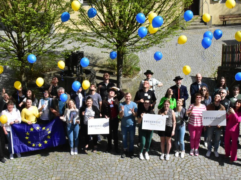 Eine fröhliche Gruppe präsentiert ein Schild, während sie bunte Ballons in der Hand halten, was Gemeinschaft symbolisiert.